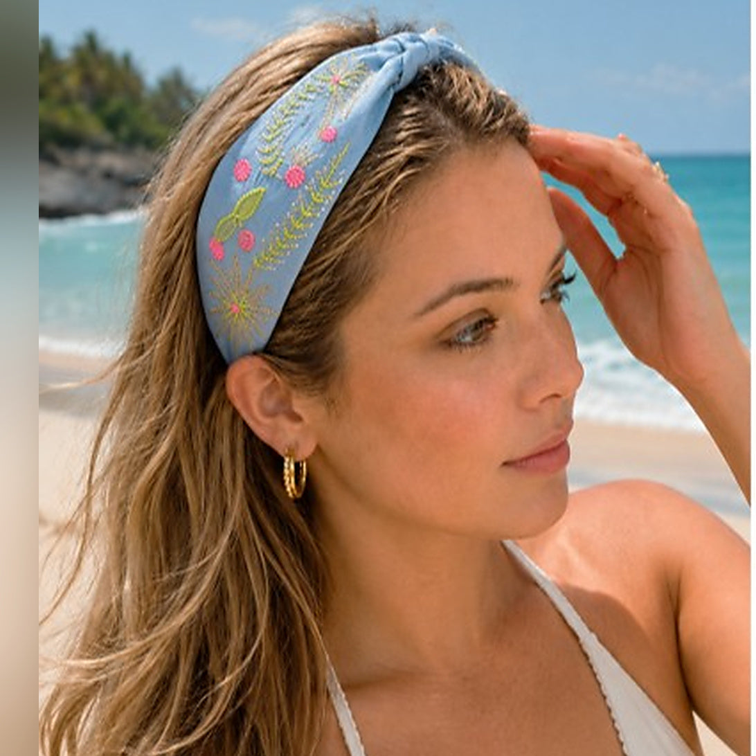 A woman on a beach wearing a light blue fabric headband with embroidered floral details in pink, yellow, and green. She is wearing a white halter top and gold hoop earrings, with the ocean and shoreline visible in the background.